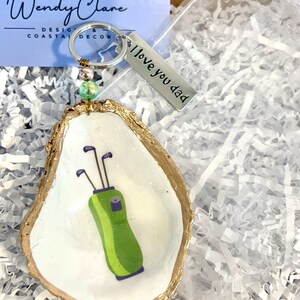 May include: A white oyster shell with a gold rim, painted with a green golf bag and clubs. The shell is attached to a silver keychain with a charm that reads "I love you dad".