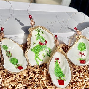 Peut inclure: Quatre décorations de Noël en forme d'huître avec des bordures dorées. Chaque décoration présente un motif différent d'arbre de Noël vert peint sur un fond blanc. Les décorations sont présentées dans une boîte remplie de papier d'emballage brun.