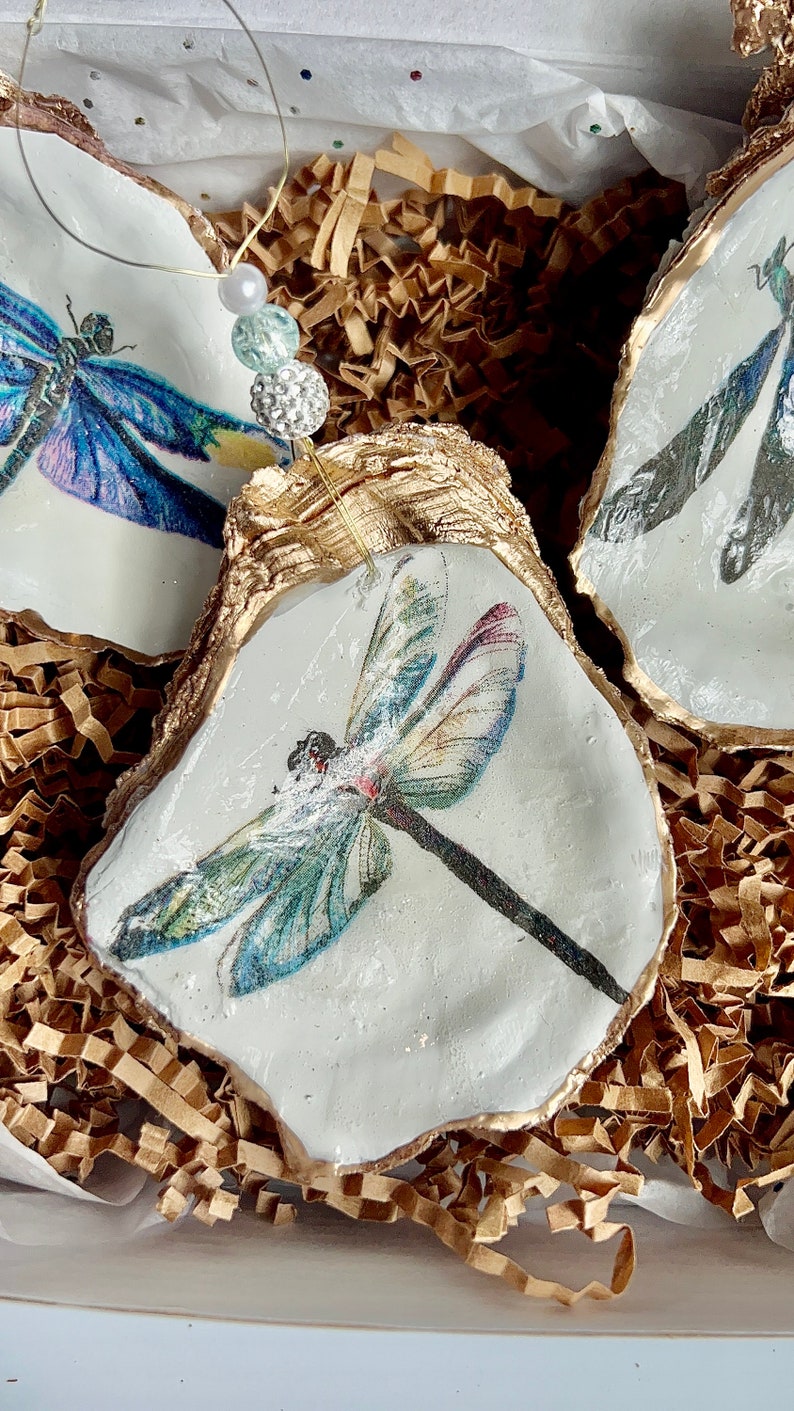 May include: Three white oyster shell ornaments with gold trim, each featuring a different dragonfly design. The dragonflies are painted in shades of blue, green, and purple.