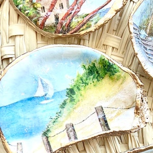 May include: Three painted oyster shells with gold trim. The first shell depicts a red-roofed building with palm trees. The second shell shows a sailboat on a blue body of water with a sandy beach and a wooden fence. The third shell shows a beach scene with a lighthouse in the distance.