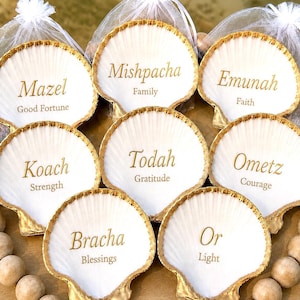 Peut inclure: Un ensemble de huit coquilles Saint-Jacques blanches aux bords peints en or, chacune portant un mot hébreu et sa traduction en anglais en lettres dorées, incluant Mazel, Mishpacha, Emunah, Koach, Todah, Ometz, Bracha et Or, disposées en cercle.