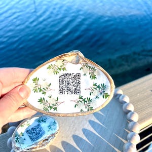 May include: Two seashell trinket dishes with floral designs and QR codes. One is held by a hand, the other rests on a wooden tray with a beaded border. The shells have a gold trim and are set against a blurred water background.