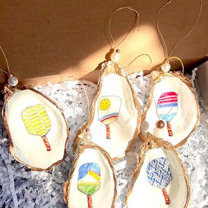 May include: Five oyster shell ornaments with gold trim, each featuring a different hand-painted design of a paddle. The ornaments are displayed in a box with white shredded paper. The text on the sticker reads "Thank you for supporting my small business."