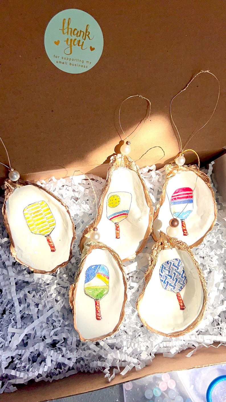 May include: Five oyster shell ornaments with gold trim, each featuring a different hand-painted design of a paddle. The ornaments are displayed in a box with white shredded paper. The text on the sticker reads "Thank you for supporting my small business."
