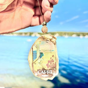 May include: An oyster shell ornament featuring a scenic image of Capri, Italy, with the word "CAPRI" printed at the bottom. The shell is suspended by a gold-colored string with two decorative beads. The background is a blurred view of the ocean and sky.