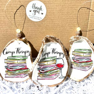 May include: Three gold oyster shell ornaments with a white background. Each ornament features a stack of colorful books, with the text "Group Therapy" written above the books. One ornament also includes a glass of red wine.