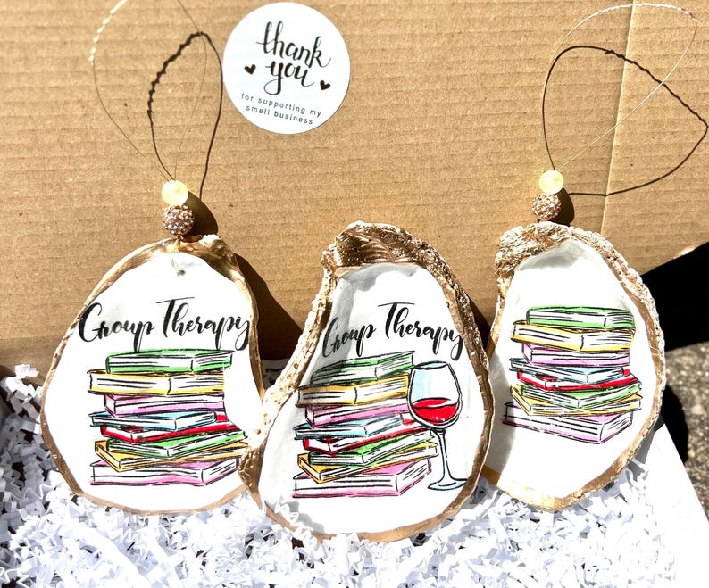 May include: Three gold oyster shell ornaments with a white background. Each ornament features a stack of colorful books, with the text "Group Therapy" written above the books. One ornament also includes a glass of red wine.