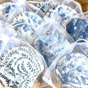 May include: A collection of small, decorative ceramic shells with blue and white floral patterns, packaged in individual white organza bags. The shells are perfect for holding small trinkets or jewelry.