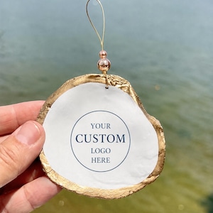Könnte beinhalten: Ein dekoratives Austernschalen-Ornament mit goldbemaltem Rand und einer weißen Mitte, die den Text YOUR CUSTOM LOGO HERE trägt. Das Ornament verfügt über eine goldene Drahtschlaufe zum Aufhängen, verziert mit zwei kleinen, runden, metallisch rosa Perlen.