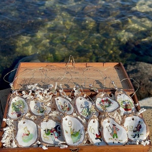 May include: A wooden box filled with 12 oyster shell Christmas ornaments. Each ornament is hand-painted with a different scene and number. The box is labeled "Stewart's Duplex Safety Pins".