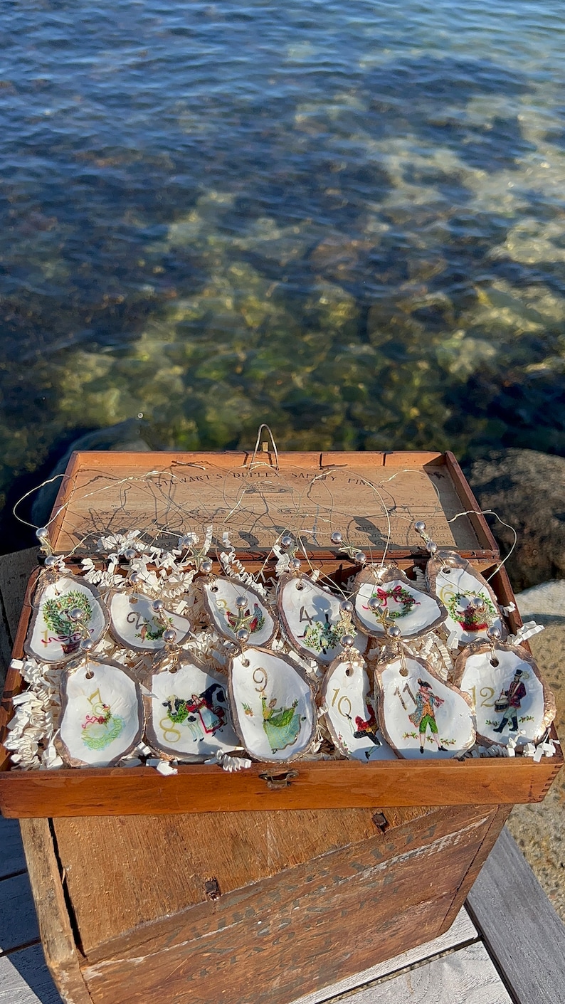 May include: A wooden box filled with 12 oyster shell Christmas ornaments. Each ornament is hand-painted with a different scene and number. The box is labeled "Stewart's Duplex Safety Pins".