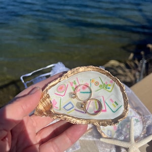 May include: A gold-edged oyster shell ring holder with a colorful patterned interior. Two gold rings with gemstones rest inside. A starfish and the ocean are in the background.