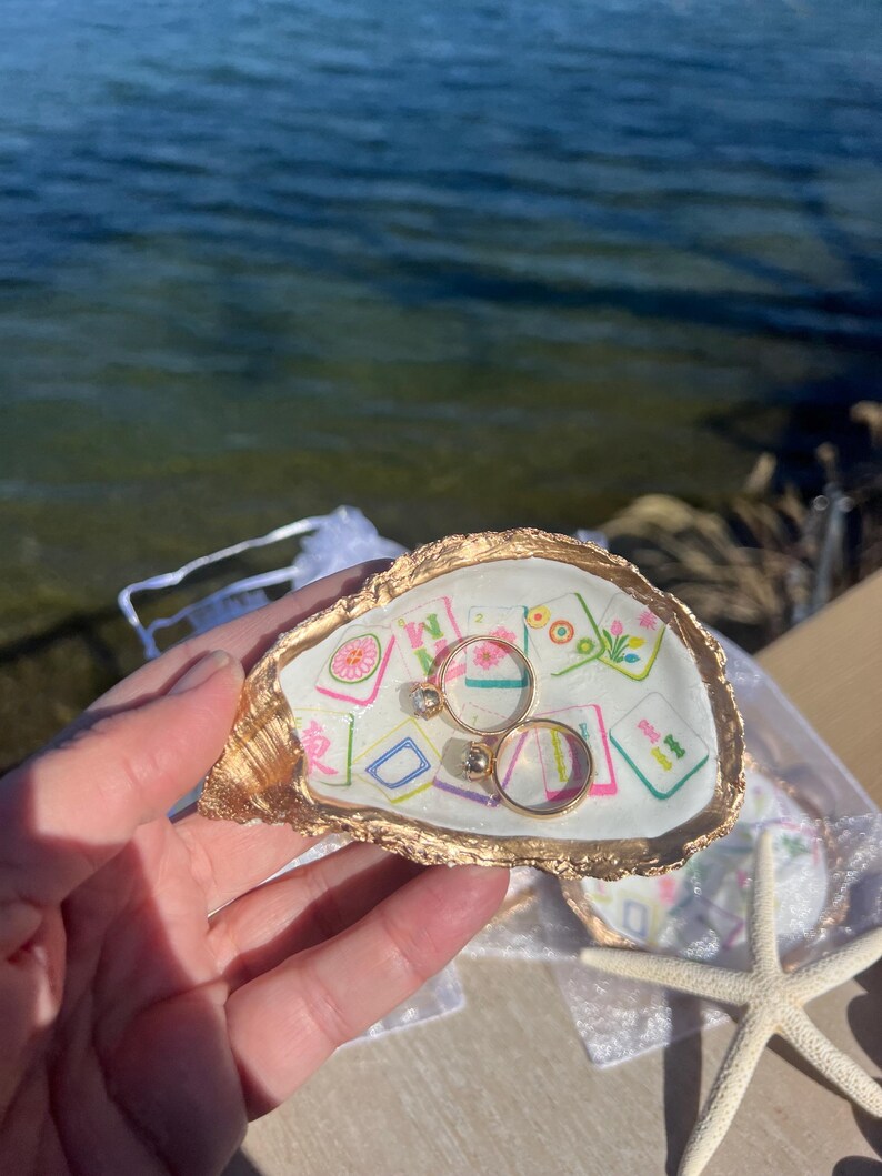 May include: A gold-edged oyster shell ring holder with a colorful patterned interior. Two gold rings with gemstones rest inside. A starfish and the ocean are in the background.