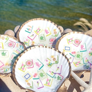 May include: Four white scallop shells with gold rims, decorated with colorful Mahjong tile designs. The shells are arranged on a wooden surface near the water, with a starfish visible. The tiles feature various symbols and characters.