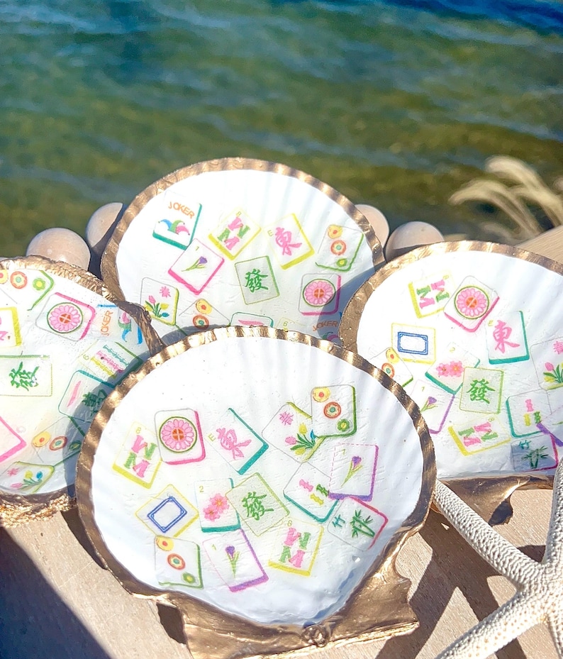 May include: Four white scallop shells with gold rims, decorated with colorful Mahjong tile designs. The shells are arranged on a wooden surface near the water, with a starfish visible. The tiles feature various symbols and characters.