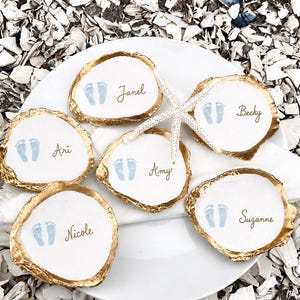 May include: Five white oyster shells with gold-painted edges, each featuring a pair of light blue baby footprints and a name written in gold script. The names are Janel, Becky, Ari, Amy, and Nicole. A white starfish rests across the center shells.