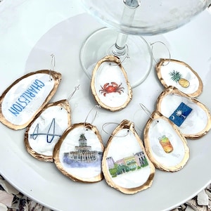 May include: A collection of oyster shell wine charms with gold edges and various watercolor designs. The charms feature images of a crab, pineapple, Charleston landmarks, and a cocktail. They are arranged on a white plate with a wine glass.
