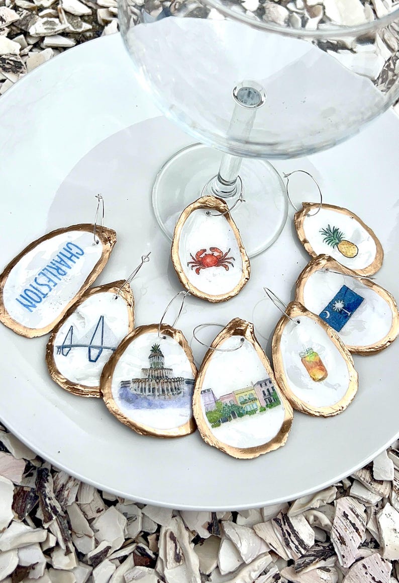 May include: A collection of oyster shell wine charms with gold edges and various watercolor designs. The charms feature images of a crab, pineapple, Charleston landmarks, and the South Carolina flag. They are arranged on a white plate.