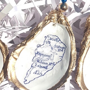 May include: Hand-painted oyster shell ornament with a gold-painted edge. The white interior features a blue map of Hilton Head Island. A blue and gold bead adorns the top, attached by a thin wire. The ornament is displayed on white paper strips.