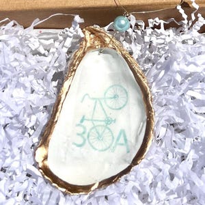 May include: An oyster shell ornament with a gold-painted edge. The interior features a light blue bicycle illustration and the text "30A". A light blue bead hangs from the top. The ornament is set against white paper filler.