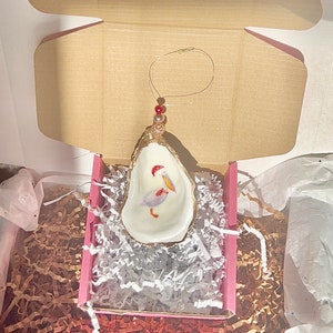 May include: A white oyster shell ornament with a gold rim, featuring a painted pelican with a red beak and a red and gold Christmas ornament hanging from the top.