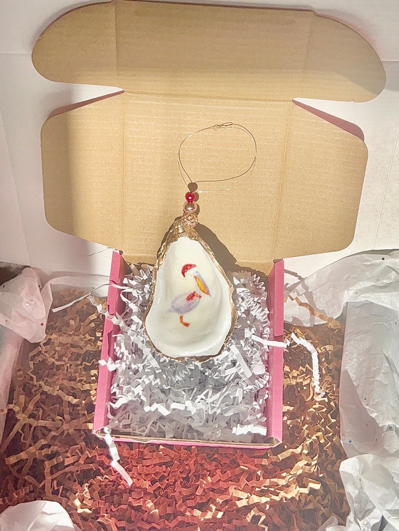 May include: A white oyster shell ornament with a gold rim, featuring a painted pelican with a red beak and a red and gold Christmas ornament hanging from the top.