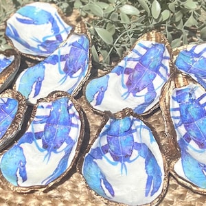 K&ouml;nnte beinhalten: Ein Satz von acht Austernmuscheln, die mit blauen Hummern bemalt sind. Die Muscheln sind in einem Kreis auf einer gewebten Oberfl&auml;che angeordnet.