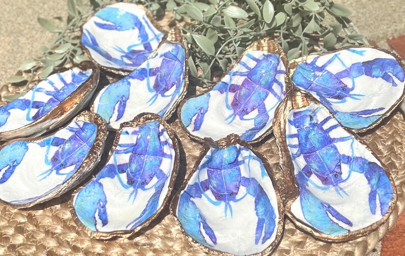 K&ouml;nnte beinhalten: Ein Satz von acht Austernmuscheln, die mit blauen Hummern bemalt sind. Die Muscheln sind in einem Kreis auf einer gewebten Oberfl&auml;che angeordnet.