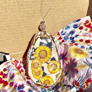 May include: A gold-painted oyster shell ornament with a floral design featuring sunflowers. The ornament is hanging from a thin gold string and has a small gold bead at the top.