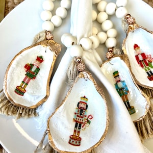 Puede incluir: Cuatro servilleteros de concha de ostra con borde dorado y diseño de cascanueces pintado. Los servilleteros están unidos a una hebra de cuentas blancas y una borla de yute. Los servilleteros están sobre un plato blanco con una servilleta blanca.