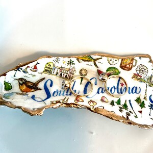 Op de afbeelding: Een witte keramische ringenschotel in de vorm van een schelp met een gouden rand. De schotel is versierd met een kleurrijke illustratie van bezienswaardigheden in South Carolina en de tekst "South Carolina". Twee gouden ringen liggen op de schotel.