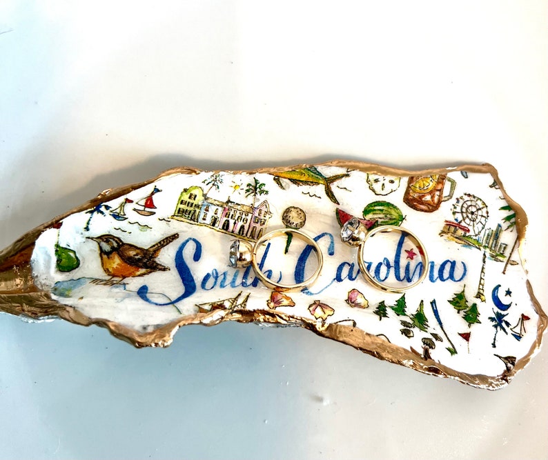 Op de afbeelding: Een witte keramische ringenschotel in de vorm van een schelp met een gouden rand. De schotel is versierd met een kleurrijke illustratie van bezienswaardigheden in South Carolina en de tekst "South Carolina". Twee gouden ringen liggen op de schotel.