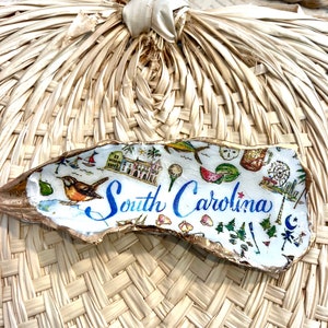 Op de afbeelding: Een witte oesterschelp met gouden accenten, beschilderd met een kleurrijke illustratie van bezienswaardigheden in South Carolina en de tekst "South Carolina".