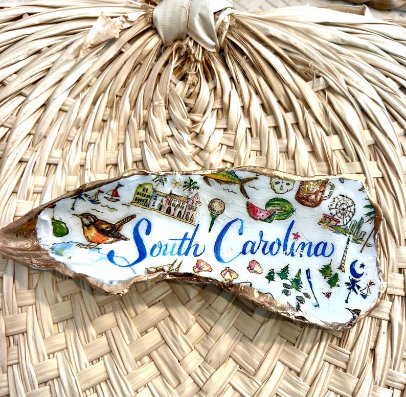 Op de afbeelding: Een witte oesterschelp met gouden accenten, beschilderd met een kleurrijke illustratie van bezienswaardigheden in South Carolina en de tekst "South Carolina".