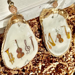 May include: Two gold-painted oyster shell ornaments with musical instrument designs. One ornament features a trumpet, bass, treble clef, and a flute. The other ornament features a saxophone, violin, harp, and a guitar.