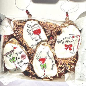 May include: Four oyster shell ornaments with gold accents and painted designs. The ornaments feature a glass of wine, a martini glass, a margarita glass, and a glass of champagne. The ornaments are decorated with red, green, and white paint. The text on the ornaments reads "Ho Ho Ho Pour the Merlot", "Making Spirits Bright", "Have a cup of cheer", and "To Do Eat Drink B-Merry".