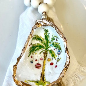 May include: A white oyster shell with a gold rim, painted with a green palm tree and colorful ornaments hanging from the branches. The shell is attached to a natural fiber tassel and a white bead garland.