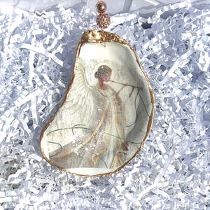 May include: A gold-trimmed oyster shell ornament with a painted image of an angel playing a flute. The ornament is decorated with glitter and has a rose gold string for hanging.