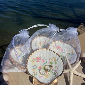 May include: A collection of seashell ring dishes with gold trim, each adorned with a colorful Mahjong tile design. Two gold rings rest in one dish. A starfish and small white bags filled with similar dishes are also present.