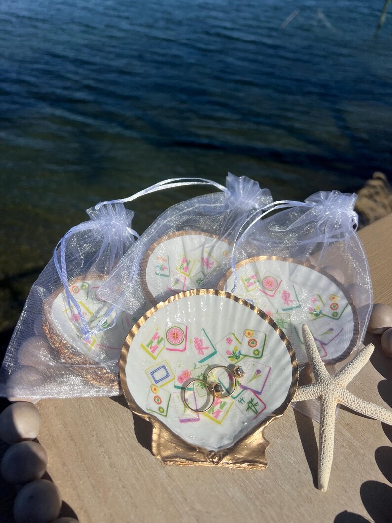 May include: A collection of seashell ring dishes with gold trim, each adorned with a colorful Mahjong tile design. Two gold rings rest in one dish. A starfish and small white bags filled with similar dishes are also present.