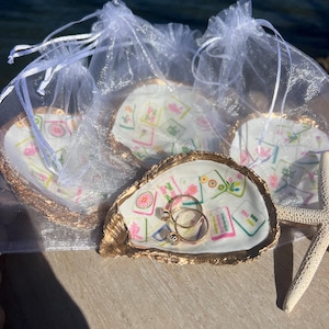 May include: Oyster shell ring dishes with gold trim, each holding a printed design and packaged in a sheer white bag. One dish holds two gold rings with clear stones. A starfish rests nearby.
