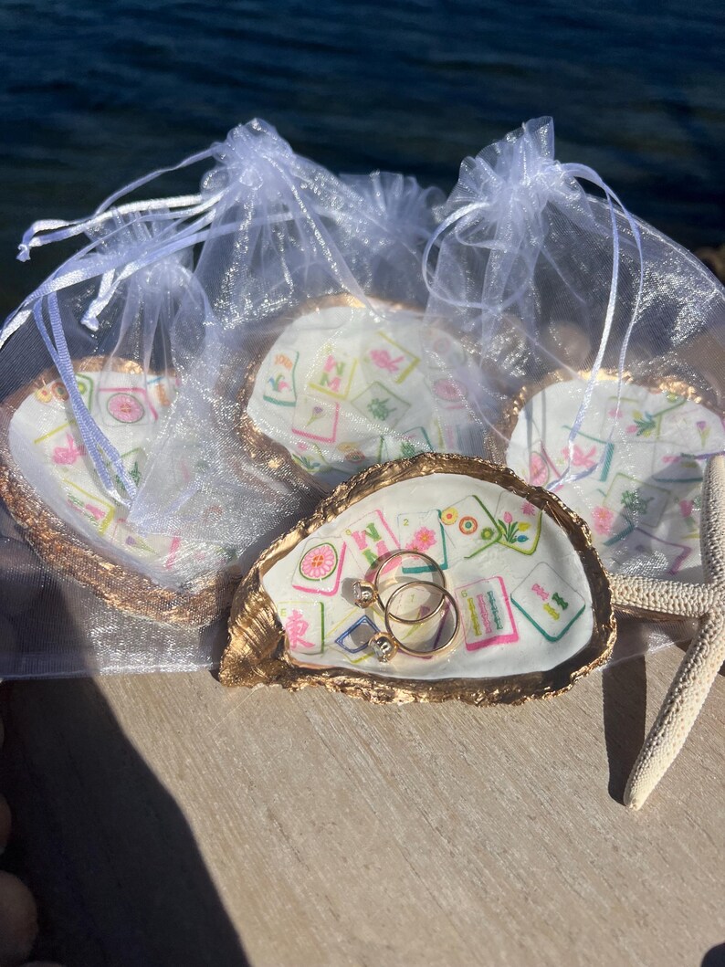 May include: Oyster shell ring dishes with gold trim, each holding a printed design and packaged in a sheer white bag. One dish holds two gold rings with clear stones. A starfish rests nearby.