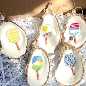 May include: Five oyster shell ornaments with hand-painted paddleball rackets. Each ornament features a different color and pattern on the racket. The ornaments are strung with gold thread and have a pearl bead at the top.