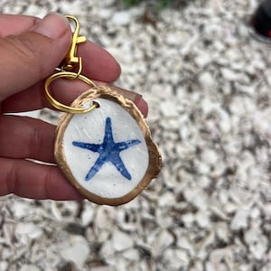 May include: A handmade keychain featuring a white, oval-shaped charm with a blue starfish design. The charm has a gold-colored border and is attached to a gold-tone keyring and clasp. The keychain is a nautical-themed accessory.