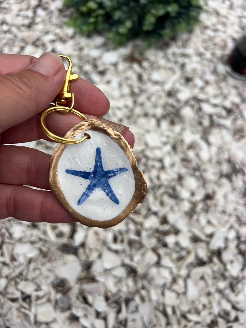 May include: A handmade keychain featuring a white, oval-shaped charm with a blue starfish design. The charm has a gold-colored border and is attached to a gold-tone keyring and clasp. The keychain is a nautical-themed accessory.