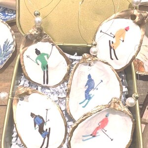 May include: Five oyster shell ornaments with gold trim, each featuring a different person skiing in colorful clothing. The ornaments are hanging from gold wire with white pearl beads.