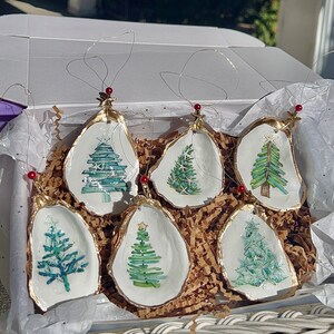 May include: Six hand-painted oyster shell Christmas ornaments with a gold trim. Each ornament features a different watercolor design of a Christmas tree.