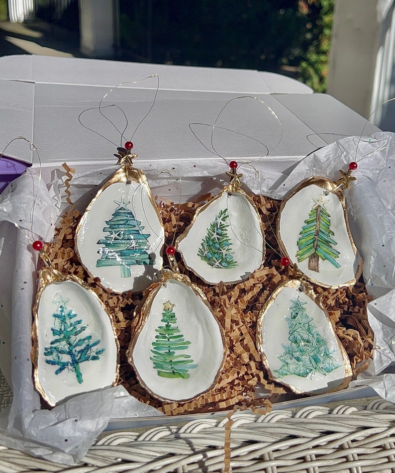 May include: Six hand-painted oyster shell Christmas ornaments with a gold trim. Each ornament features a different watercolor design of a Christmas tree.