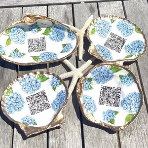 May include: Four seashell-shaped dishes with gold rims and white interiors. Each dish features a blue hydrangea floral pattern and a QR code. Two starfish are placed between the dishes on a wooden surface.