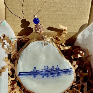 Może przedstawiać: Biała ceramiczna ozdoba w kształcie muszli ostrygi z pozłacanym brzegiem. Ozdoba przedstawia niebiesko-białą ilustrację załogi wioślarskiej w łodzi. Ozdoba jest zawieszona na złotym łańcuszku z niebiesko-złotą koralikiem.